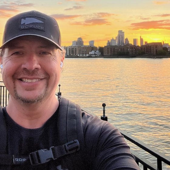 Jeff smiling on the Thames riverside at sunset, wearing a GORUCK cap and rucksack with the sternum strap clipped, the City skyline behind
